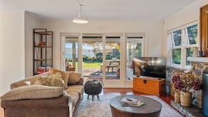 A living room with a sofa, ottoman, coffee table, TV, bookshelf, and a view of the outdoors. - Waterside Bliss - Taupo Holiday Home (Taupo)