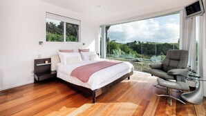 Une chambre à coucher moderne avec un grand lit, une fenêtre fermée par une paroi de verre et un fauteuil gris.