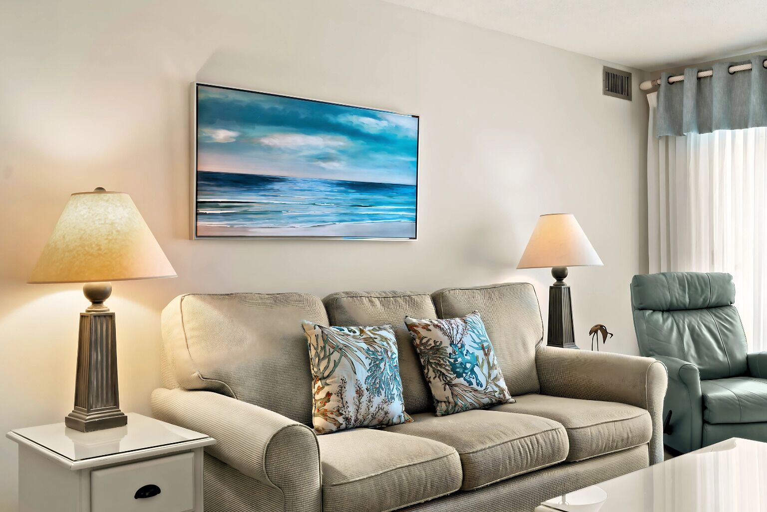 A living room with a beige sofa, two decorative pillows, a side table, a lamp, a chair, and a painting of a beach scene.