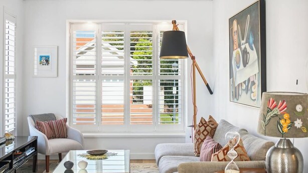 A living room with a sofa, armchair, coffee table, and a large window with shutters.