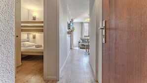 Un couloir d’hôtel avec un plancher en bois, une chambre avec des lits superposés sur la gauche, un coin salon avec une table et des chaises, et une porte en bois sur la droite.