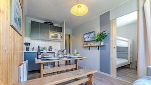 Un espace de vie compact comprenant un coin cuisine, un espace repas avec un banc en bois, un lit et un téléviseur fixé au mur.