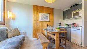 Un salon chaleureux avec un banc en bois, un canapé et une table à manger pour deux personnes. On aperçoit, au fond, une cuisine équipée d’appareils électroménagers modernes.