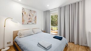 Ein modernes Schlafzimmer mit Bett, Nachttischlampe, einem Bild und Blick auf Grün durch das Fenster.