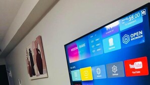 Een moderne woonkamer met een tv aan de muur waarop diverse apps worden weergegeven, een kleine tafel met stoelen en een plant.