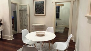 Un coin repas avec une table ronde, des chaises blanches et un bol posé sur la table. Un tableau encadré orne le mur, et une petite table d’appoint se trouve à proximité.