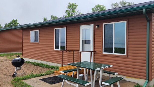 A single-story cabin with a green roof, a front door, and a small porch with a table and benches. - 3-bedroom Cabin at West Wind Resort in beautiful Waskish, MN on Upper Red Lake. (Waskish)