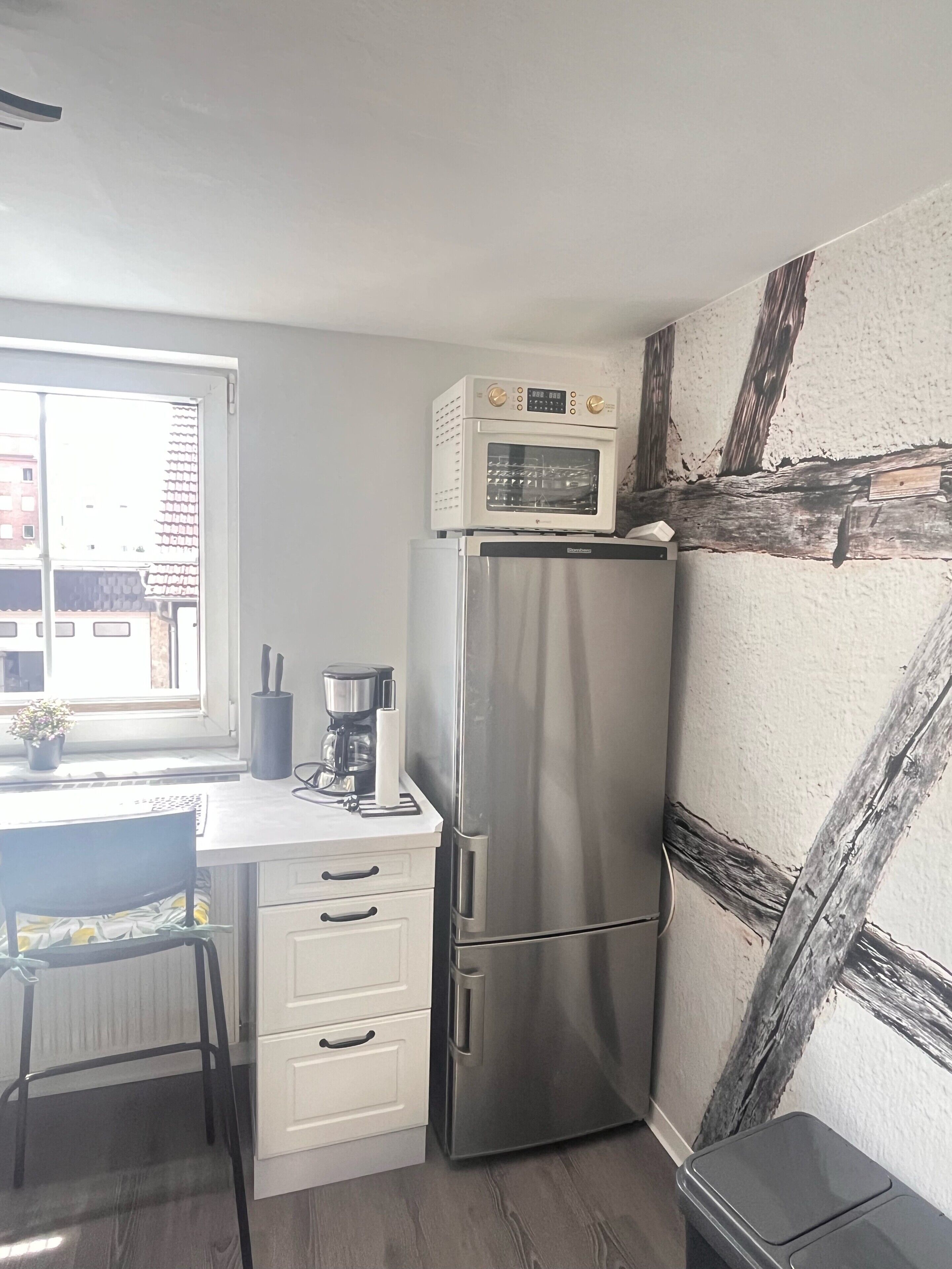 Una cocina compacta con un refrigerador de acero inoxidable, un microondas arriba, una cafetera y una ventana con vista a edificios.