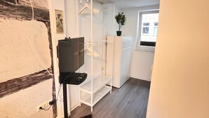Et værelse med trægulv, hvide vægge, et hvidt reolsystem, en sort stol og et vindue med udsigt til bygninger.