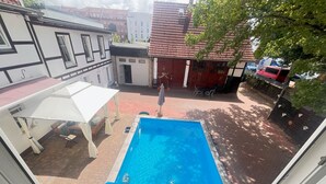 Udsigt til en swimmingpool fra et vindue, med et overdækket udendørs opholdsområde og en rød bygning i baggrunden.