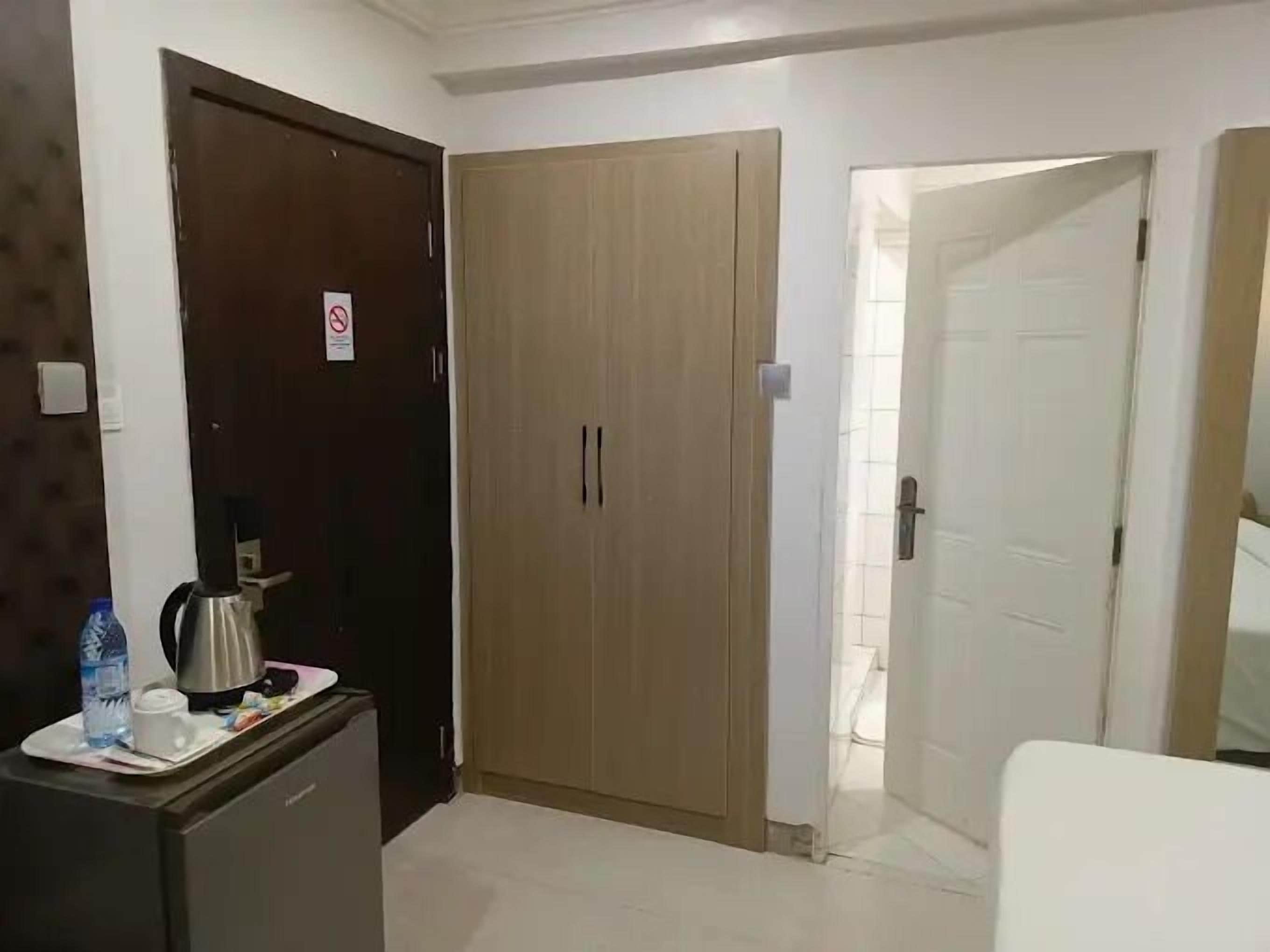Une chambre d’hôtel avec une armoire en bois, un bureau avec une bouilloire et une porte blanche donnant sur une autre pièce.