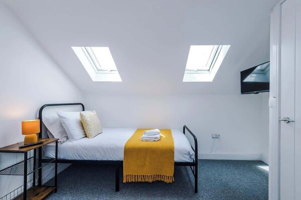 A single bed with a yellow throw blanket, a bedside table with a lamp, and a TV mounted on the wall.