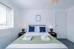 A hotel room with a bed, two bedside tables, a window with blinds, a door, and a wall-mounted picture. - Modern Snug 3BR | W/ Parking | Shrewsbury Town Gem (Shropshire)