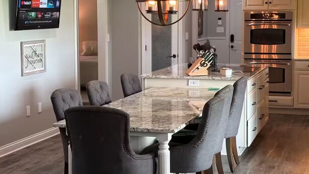 Una cocina moderna con una isla de mármol, sillas tapizadas oscuras, iluminación colgante y un televisor de pantalla plana montado en la pared.