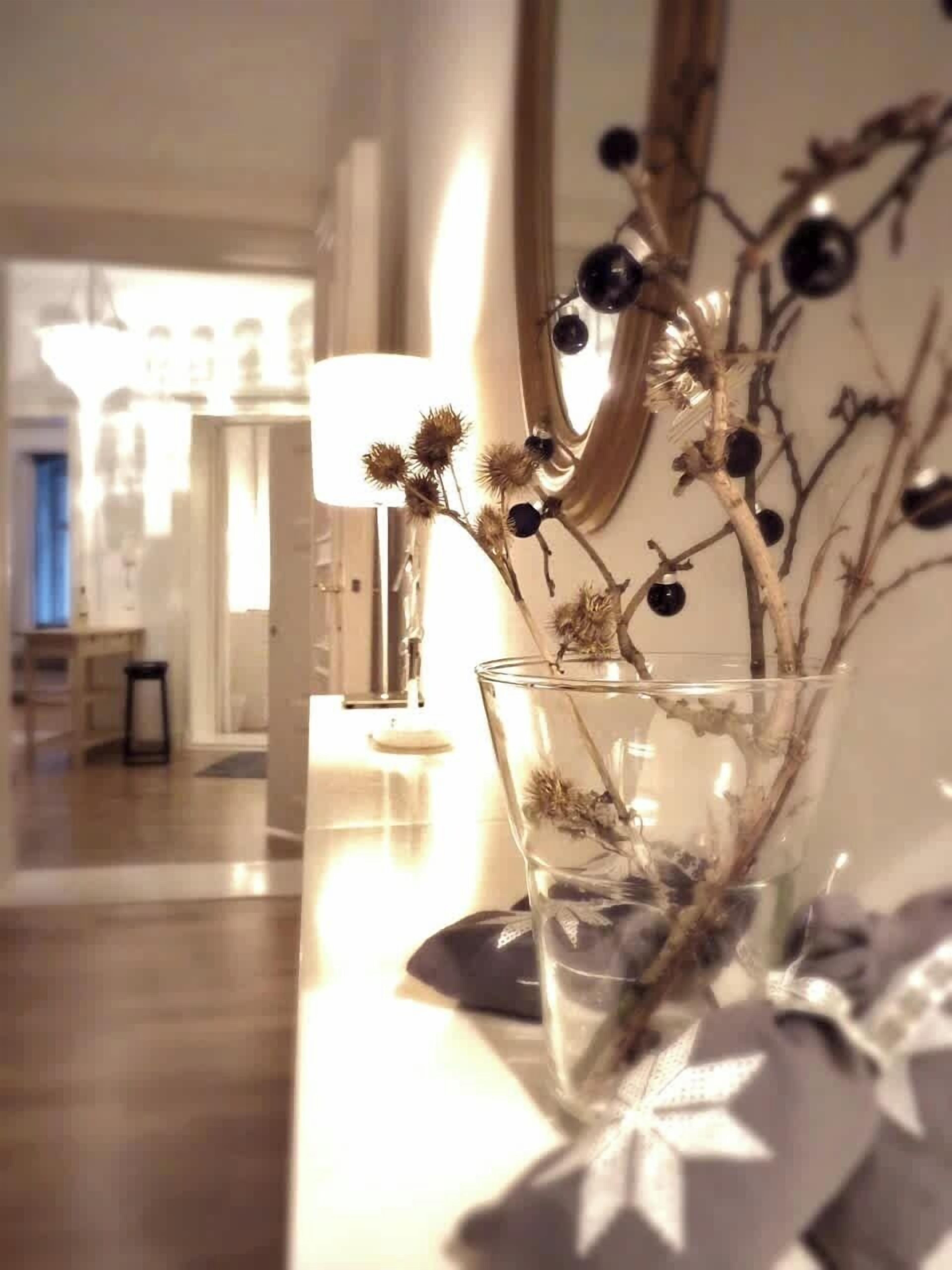 Un florero de vidrio transparente con ramas secas y bayas sobre una mesa en una habitación bien iluminada, con una lámpara y un espejo que refleja el interior de la estancia.