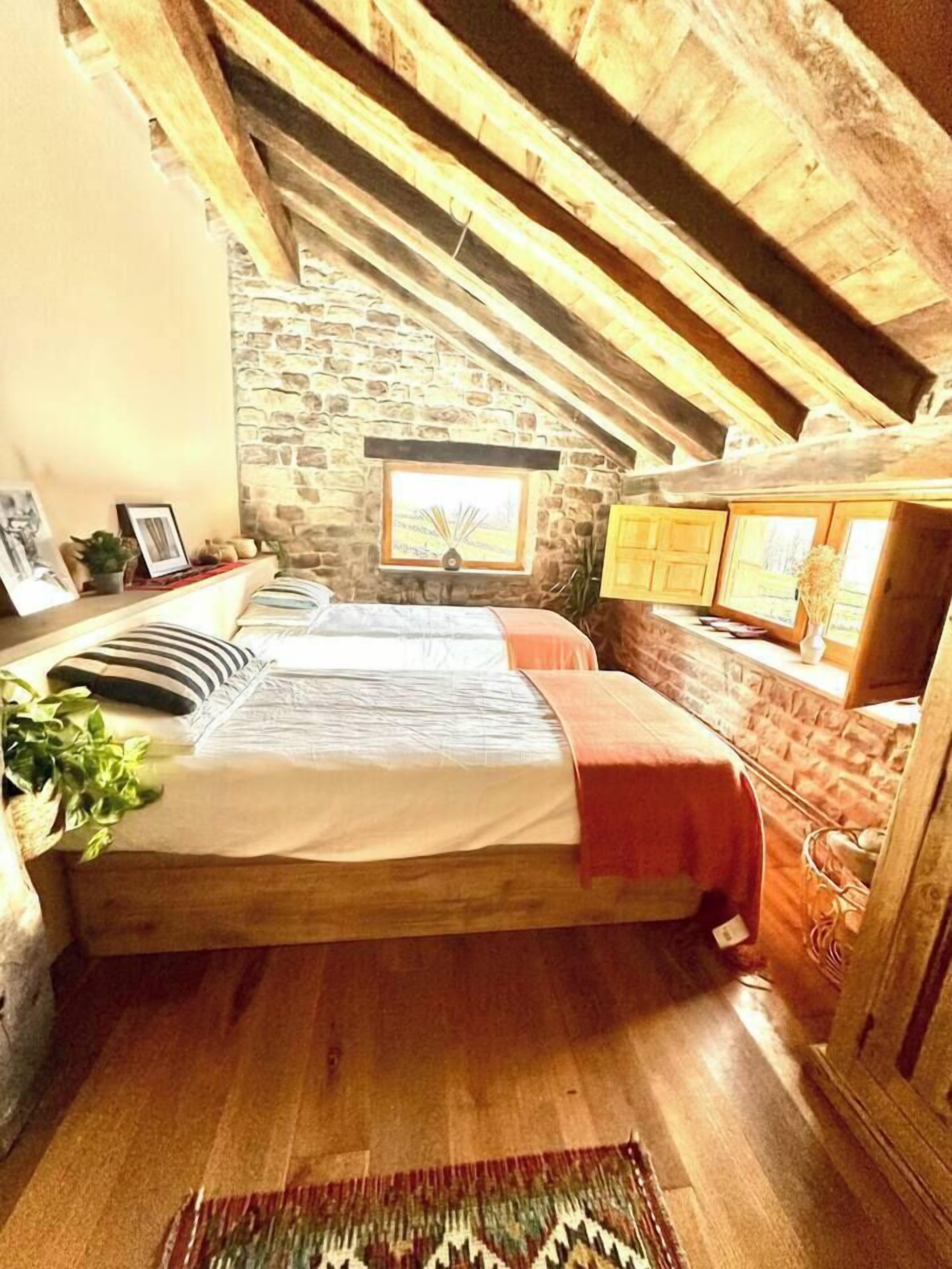 Ein gemütliches Schlafzimmer im Dachgeschoss mit Steinwand, Holzbalken, einem Bett, einem Fenster und einer kleinen Küchenzeile.