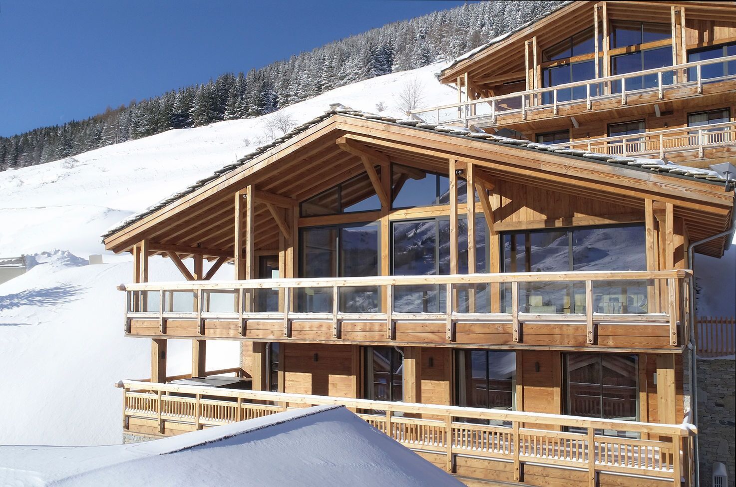 A multi-story wooden chalet with balconies, large windows, and a snowy mountain backdrop.