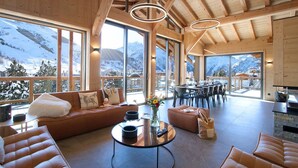 Amplia sala de estar con un gran ventanal con vistas a un paisaje montañoso nevado, vigas de madera en el techo y una zona de comedor con mesa y sillas.
