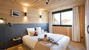 Un dormitorio con techo de madera, cama con ropa de cama blanca, un osito de peluche y una ventana con cortinas.