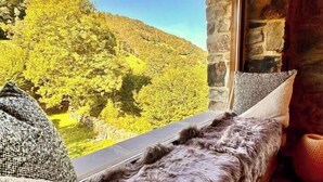 A cozy room with a wooden ceiling, stone walls, and a large window offering a view of a green hillside.
