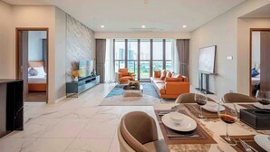A modern living room with a sofa set, a dining table with chairs, a TV, and a large window with a view of buildings. - Luxury Apartment Hotel - The Metropole (Ho Chi Minh City)
