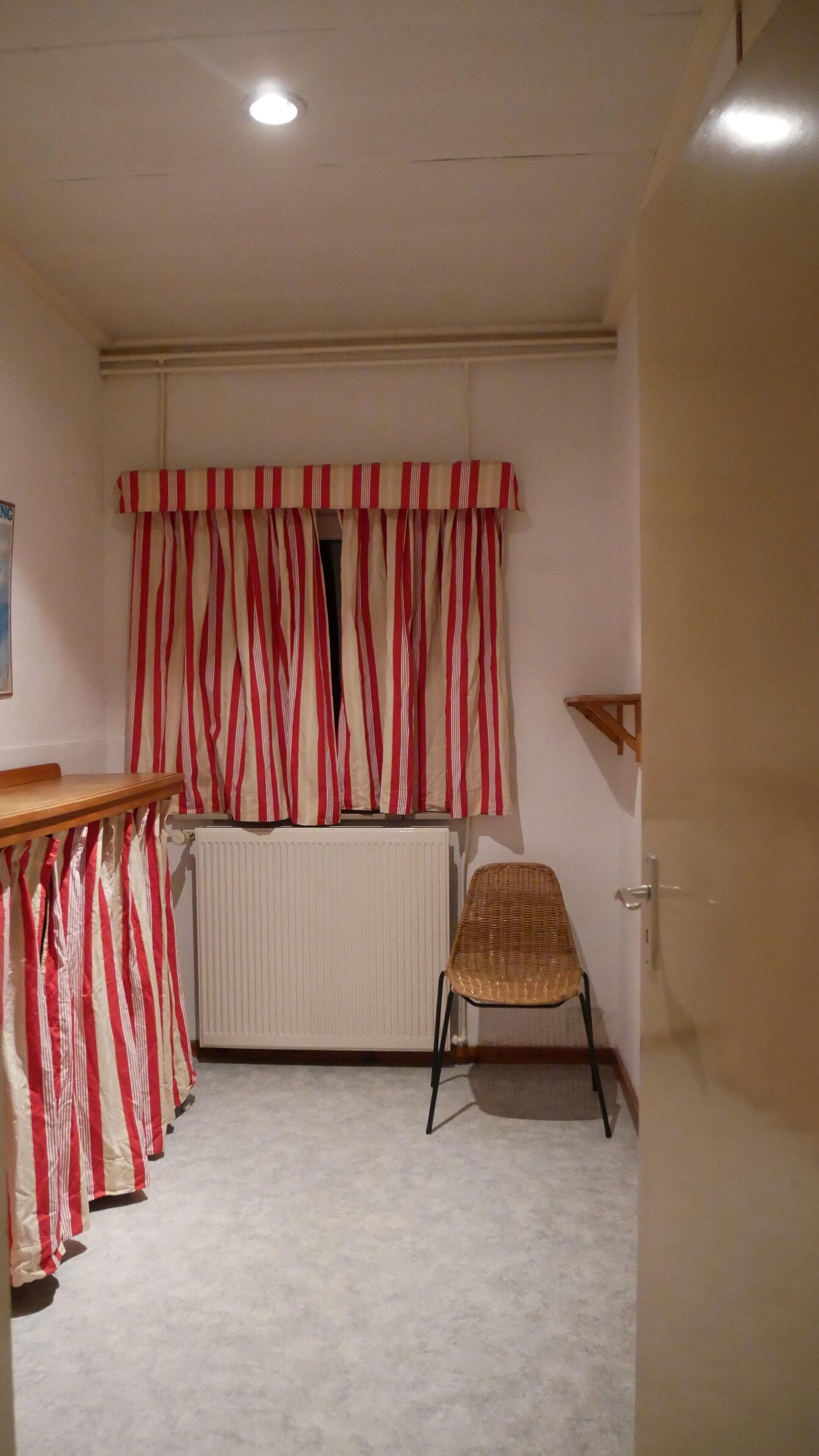 Una stanza con un termosifone, una sedia in vimini, una tenda a righe e un tavolo di legno.