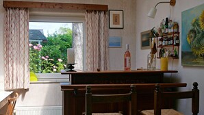 A room with a wooden bar, two chairs, a window with curtains, a radiator, and a shelf with bottles. - Cozy Family Home With Lovely Garden (Kamperland)