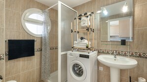 Ein kleines Badezimmer mit Waschmaschine, Waschbecken, Dusche mit Glastür und rundem Spiegel.
