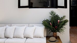 A white sofa with white cushions, a wooden side table with a hat, and a potted plant with green leaves and pink flowers. - Private Pool, Chic, Step’s from beach in beautiful Puerto Escondido - WiFi, AC (Puerto Escondido)