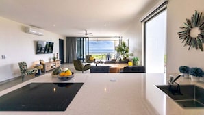 Un salón moderno con una amplia mesa central, un televisor de pantalla plana montado en la pared y vistas al océano a través de grandes ventanales.