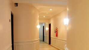 Un couloir aux murs de couleur crème, avec des lambris blancs et un sol carrelé. Des luminaires sont fixés aux murs et un extincteur est accroché au mur.
