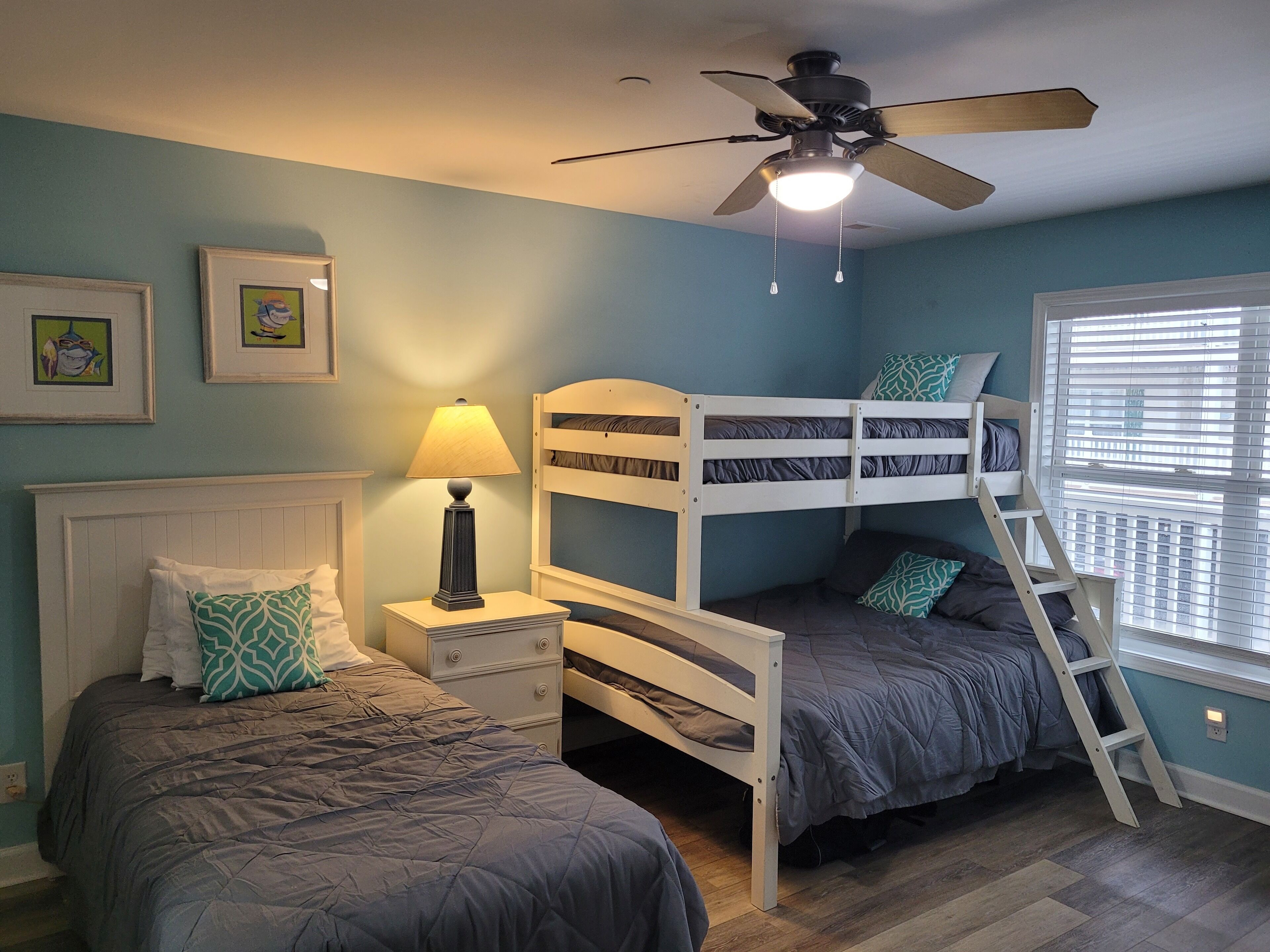 Une chambre avec des lits superposés, un ventilateur de plafond, une table de chevet avec une lampe et une fenêtre avec des stores.