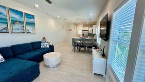 A modern living room with a blue sofa, a round ottoman, and a dining area with a table and chairs. - Docked at Dakota Sunset - Mid Island Pool Home (Fort Myers Beach)