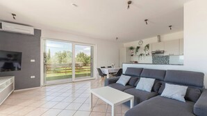 Ein modernes Wohnzimmer mit einem grauen Sofa, einem weißen Couchtisch und einem Fernseher auf einem Lowboard. Ein Schiebetürfenster führt auf einen Balkon mit Blick auf Grünflächen.
