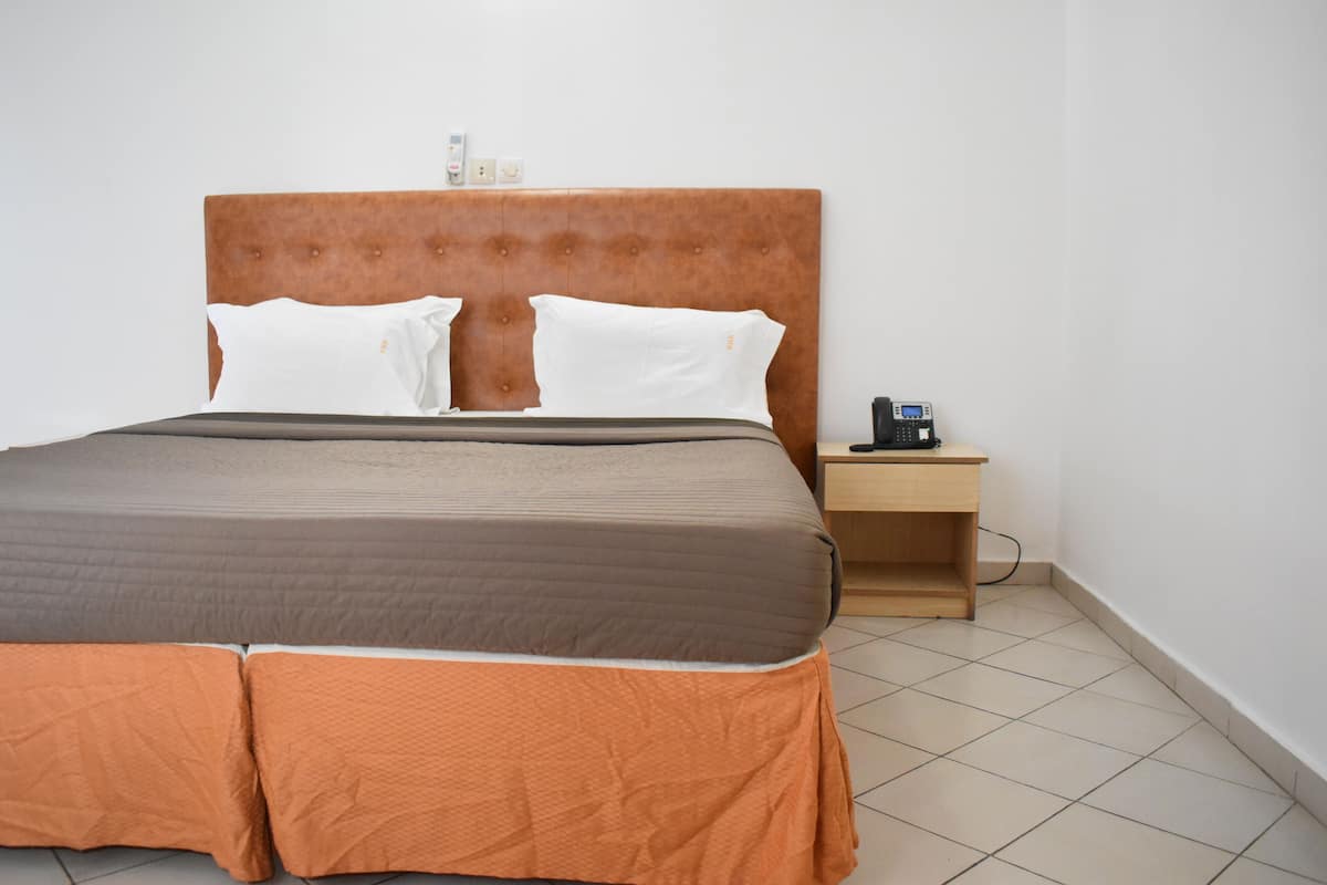 Ein ordentlich bezogenes Bett mit braunem Kopfteil, weißen Kissen und einer orangefarbenen Bettdecke. Dazu ein Nachttisch aus Holz mit einem Telefon und einer kleinen Lampe neben dem Bett.