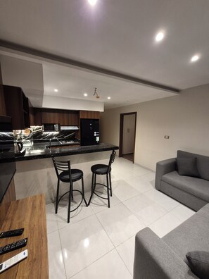 Eine moderne Küche mit einer zentralen Kochinsel, zwei Barhocker, einem grauen Sofa und einem Holztisch mit Fernbedienungen.