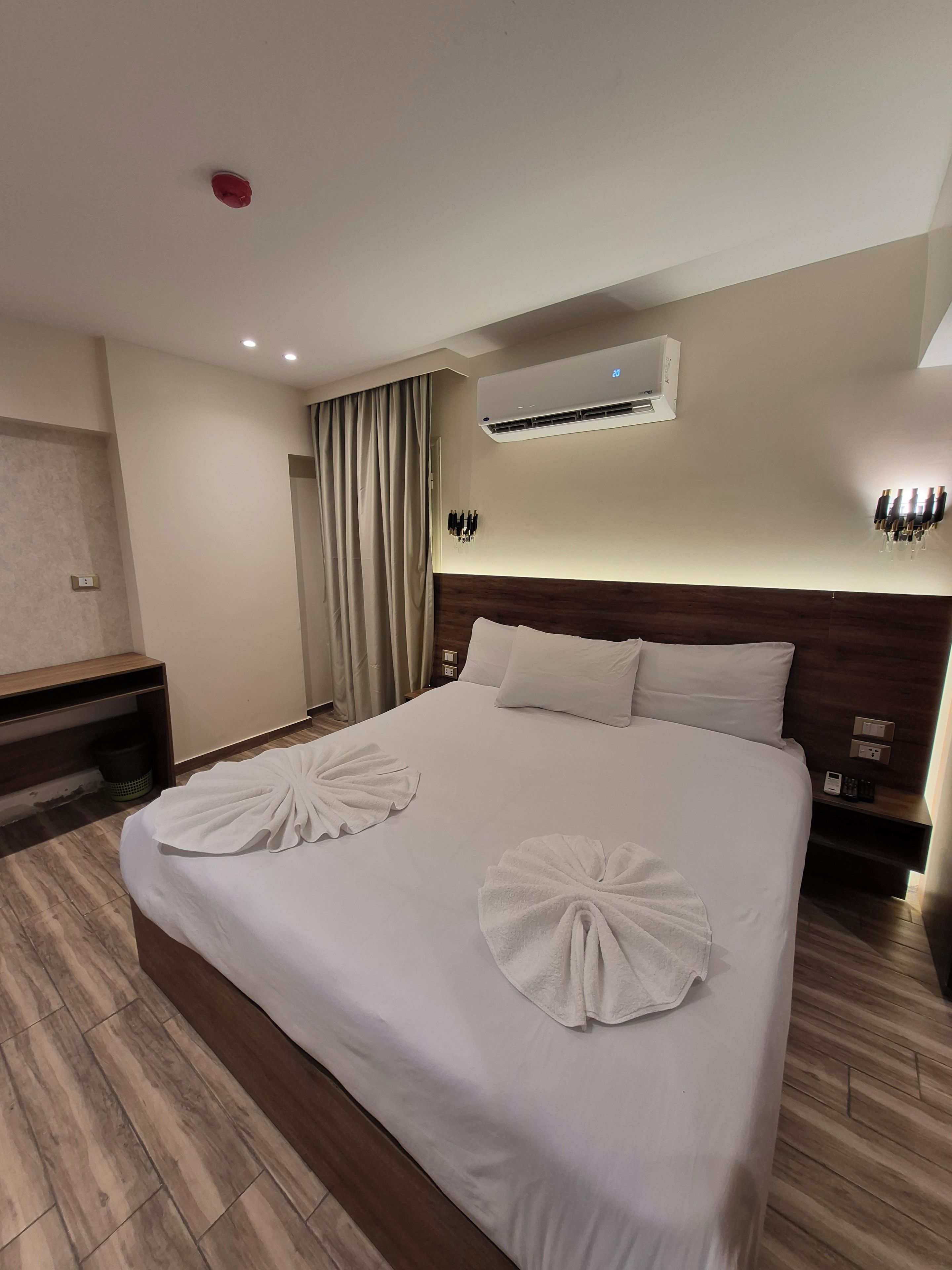 A hotel room with a large bed, two pillows, an air conditioning unit, a bedside table with a phone, and a wall-mounted light fixture.