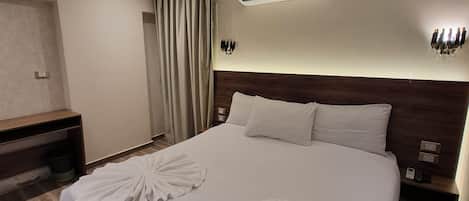 A hotel room with a large bed, two pillows, an air conditioning unit, a bedside table with a phone, and a wall-mounted light fixture.