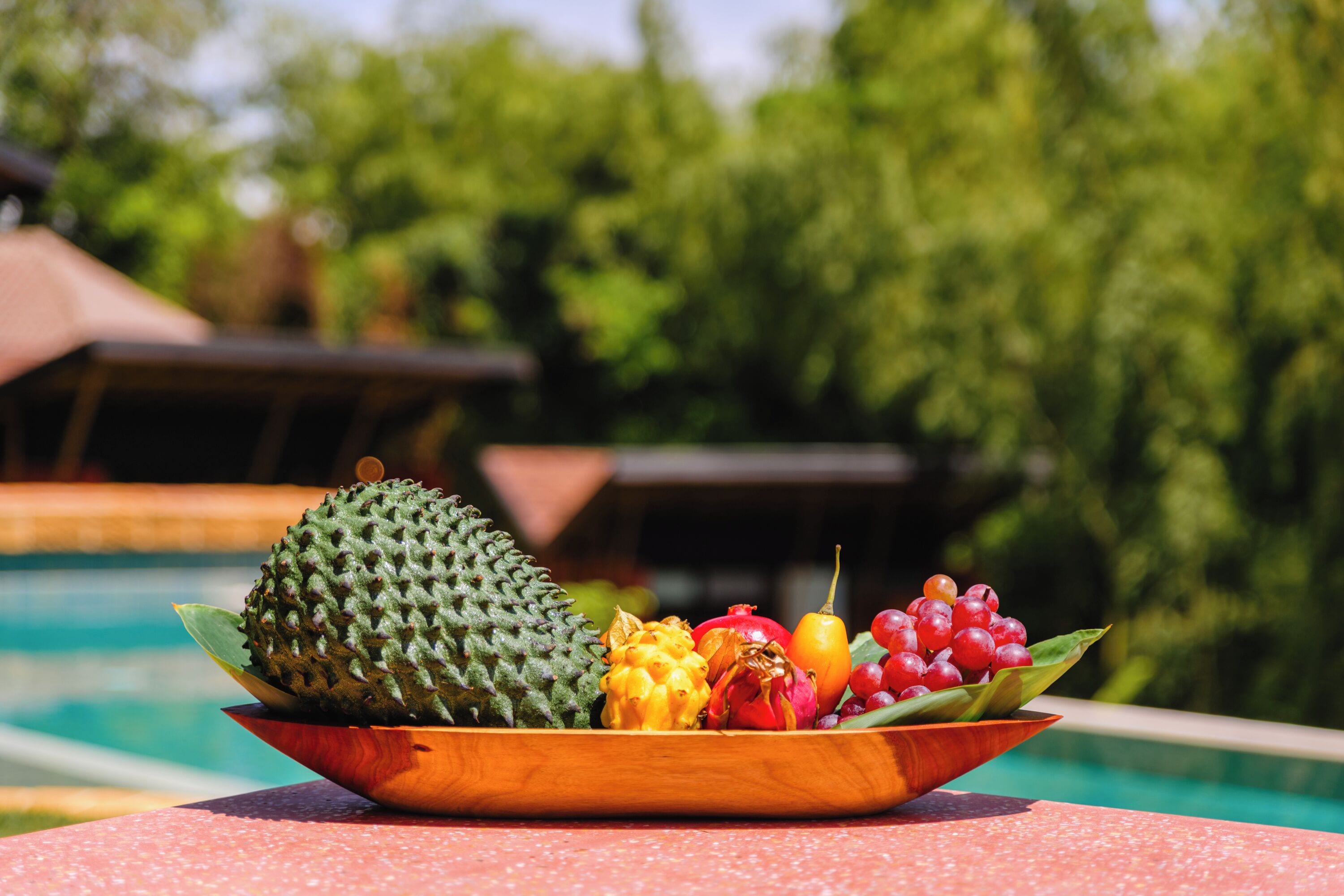 A wooden bowl filled with various fruits placed on a table by a poolside.