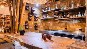 Um espaço de bar rústico com estantes de madeira cheias de garrafas, luminária de fibra natural e balcão de madeira com copos.