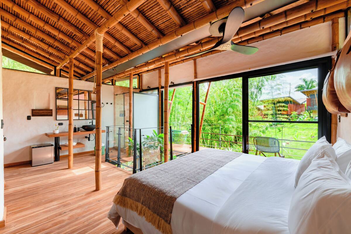 Une chambre avec un grand lit, un plafond en bois, une vue sur la verdure, une petite cuisine et un balcon.