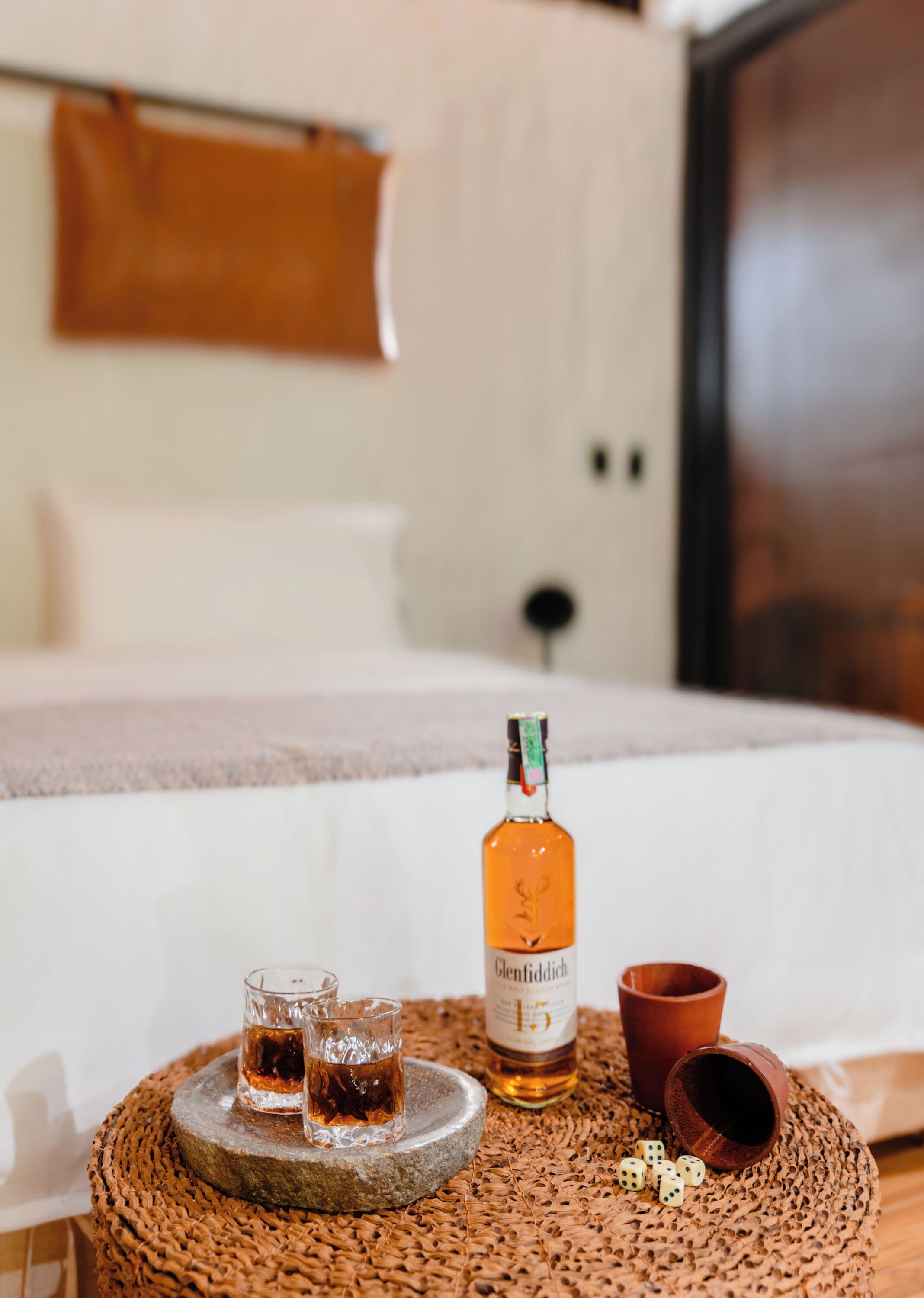 Una cama con una manta marrón, una botella de whisky Glenfiddich, dos vasos y una taza de arcilla sobre una mesa de fibras naturales.