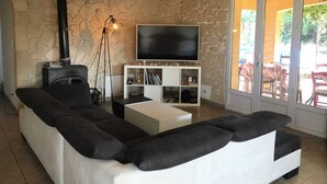 A living room with a stone wall, a black sofa, a white coffee table, a TV on a white stand, and a floor lamp. - Air-conditioned villa with large swimming pool (Le Pouget)