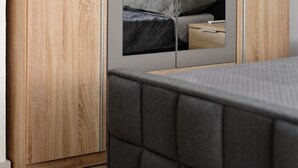 Ein Schlafzimmer mit Holzschrank, einem Bett mit grauem gepolsterten Kopfteil und einem Spiegel, der ein rotes Kunstwerk reflektiert.