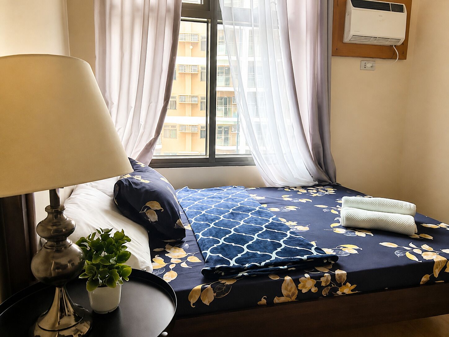 Quarto de hotel com cama, luminária de cabeceira, planta em vaso e janela com cortinas.