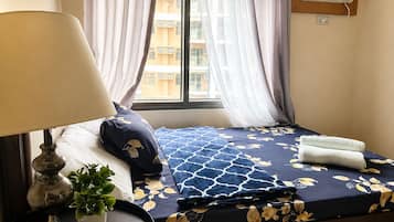 Quarto de hotel com cama, luminária de cabeceira, planta em vaso e janela com cortinas.