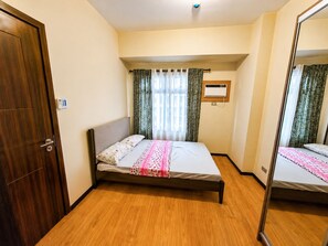 A bedroom with a wooden floor, a bed with a white and pink bedspread, a mirror, and a window with curtains. - Manila Bay View - 1BR with balcony (Pasay)