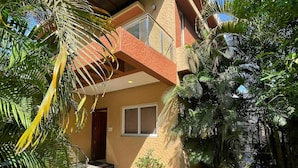 Un edificio de dos plantas con balcón, plantas en macetas y una zona de entrada con baldosas.
