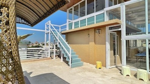 Un edificio con diseño moderno, que presenta fachada de vidrio y sección superior de madera. Hay una escalera que conduce a la entrada, y un área cubierta con estructura de techo tejida.