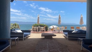 A beachside lounge with outdoor seating, umbrellas, and a view of the sea and mountains.